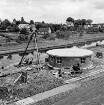 Zentralklärwerk: Anna-Heitmann-Weg: Pumpwerk in Bau: Pumphäuschen fertig, daran abgestelltes Fahrrad: Gestell mit Flaschenzug, Leiter: Betonrinne, teils offen, teils mit Abdeckung: mobile Motorpumpe, Ventil, Wasserschläuche, Rohrleitung, Baumaterialien: Bildmitte Trave: an gegenüber liegendem Ufer Weg, Gärten, Häuser, hinten Bahndamm, Signal