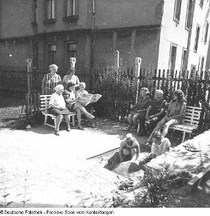 Beim Zeitungsstudium am Haus, Genrebildserie, ältere Damen auf 2 Bänken diskutieren Zeitungsmeldungen, 2 Kinder spielen im Sand