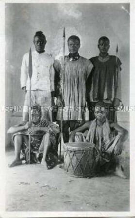 Gruppenbild von fünf Kriegern der Kaka und Jangere, mit Waffen und einer Trommel, vor neutralem Hintergrund