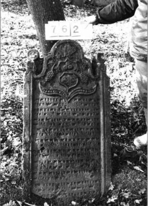 Grabstein 762 (Index: 99751585) Material: Sandstein Zustand: abgeblättert mit Textverlust Maße HxBxT: 118x62x10 cm Beschreibung: Stele, geschwungener Abschluß mit Eckakroteren, Schriftfeld eingetieft Symbolik: Trauben, Wappenschild, Ranken, Lilien, Blüten: Grabstein 762