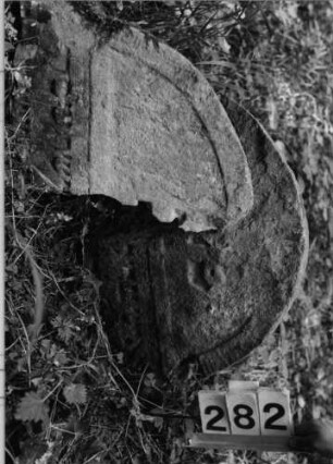 Grabstein 282 (Index: 99803423) Material: Sandstein Zustand: gespalten/eingesunk/befestigt/Textverlus Maße HxBxT: 44x68x6 cm Beschreibung: Stele mit eingezogenem Karniesbogen und profiliertem Rand, Giebelfeld: Grabstein 282