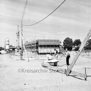 Langnese: Gewerbegebiet: Hammoorer Weg: Honigfabrikation: Betriebsgebäude in Bau: Rohbau mit Gerüsten, Baumaterialien: Baukran, Betonsilo: vorne Baugrube mit Fundament, Erdhaufen, davor Arbeiter bei Vermessung und Einrichten einer Baustelle, Planmappe auf Schubkarre, provisorische Baustromleitung: hinten weiteres Firmengabäude in Bau, Haufen Mauersteine: im Hintergrund Wohnhäuser