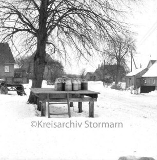 Rethwischdorf: Dorfplatz: links Baum: davor Milchkannenbock: dahinter landwirtschaftlicher Anhänger: im Hintergrund Häuser: Winter
