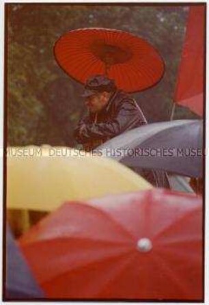 Ein Mann in schwarzer Regenkleidung hält einen japanischen Schirm über seinen Kopf, vor ihm westliche Regenschirme (Prämiertes Foto Sonderthema: Meinungsfreiheit - Ihre Chancen und Gefährdungen)