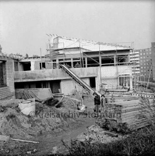 Evangelisches Gemeindehaus: Masurenweg: Rohbau: davor Erdhaufen, Bretterstapel, Baumaterial, Förderband und zwei Arbeiter: hinten rechts Hochhäuser