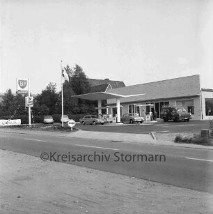 BP-Tankstelle: Hamburger Straße: Neubau: rechts Gebäude: davor Zapfsäulen mit tankenden Pkws, Kleintransporter und Personen: links BP-Schild und parkende Pkws: dahinter Bäume und Wohnhäuser