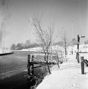Trave: zugefrorener Fluss: in der Mitte Bootsanleger hinter Bäumen: rechts Zaun und Fahnenmast: dahinter eingezäunte Weiden: hinten rechts Haus: im Hintergrund Bäume: Winter