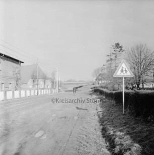 Dorfstraße: Landesstraße Richtung Reinfeld: rechts Verkehrsschild: dahinter Hecke und Garten mit Bäumen: hinten Mitte Fahrradfahrer: links Bauernhäuser: davor Zaun und Strommasten