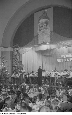 Blick vom Zuschauerraum auf die Bühne mit Orchester, Dirigenten und einem Menschen im Weihnachtsmannkostüm bei der Weihnachtsfeier des VEB Bodenbearbeitungsgeräte