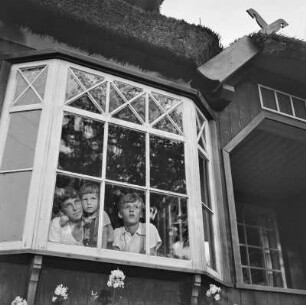 Elisabeth Gremmler schaut mit ihrem Kind und einem weiteren Jungen aus dem Fenster eines reetdeckten Hauses