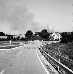 Brand: Weide in der Feldmark: vorn Straße: links Abzweigung mit Verkehrsinsel und Hinweisschild: hinten rechts Häuser und Bäume: hinten links Feld: dahinter Rauchschwaden am Himmel