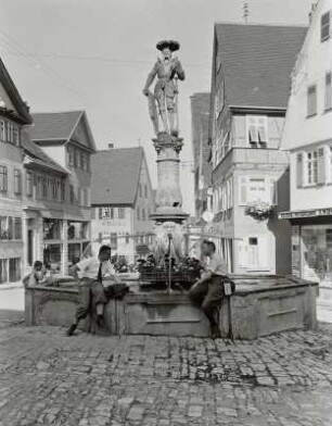 Platz mit Brunnen und zwei am Brunnenrand posierenden Wanderern