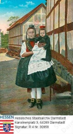 Trachten, Hessen / Frau in hessischer Tracht, ein kleines Mädchen auf dem Arm haltend, vor Fachwerkzeile stehend, Ganzfigur