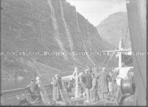 Passagiere an Deck des Hochseepassagierdampfers "München" beobachten während der Fahrt herabstürzende Wasserfälle, Nordlandreise, Norwegen