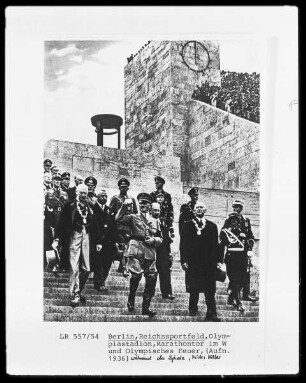 Adolf Hitler mit Gefolge im Olympiastadion während der Olympischen Spiele, im Hintergrund das Marathontor und das Olympische Feuer