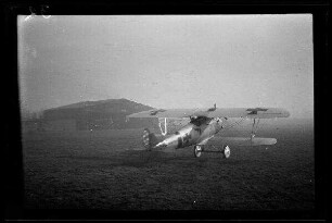 Nicht identifizierbare Ortschaft in Nordfrankreich oder Belgien: Doppeldecker mit Tatzenkreuz auf der Tragfläche und am Rumpf auf einem Feld, im Hintergrund ein Hangar ("Jagdstaffel Czermak")