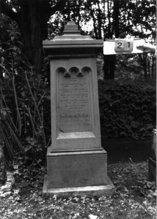 Grabstein 21 (Index: 100101744) Material: Sandstein Zustand: im Schriftfeld abgeblättert, Aufsatz fehlt Maße HxBxT: 167x53x40 cm Beschreibung: konische Stele, profilierter Sockel, Abdeckplatte mit Aufsatz, Schriftfeld eingetieft mit dreibogigem Abschluß Symbolik: gestelztes Rundbogenfries Steinmetz: A. Epple: Grabstein 21