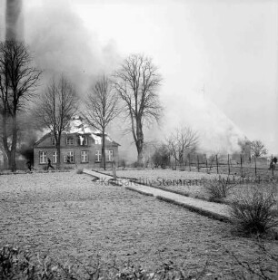Brand: Wohn- und Wirtschaftsgebäude von Landwirt Leinius: brennendes Wohnhaus hinter Baumreihe: davor rennende Feuerwehrmänner: im Vordergrund Garten