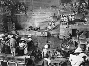 Varanasi (Benares), Indien. Touristen an Deck eines Bootes in Korbmöbeln sitzend, beobachten das Geschehen einer öffentlichen Hinduverbrennung an einem Ghat am Ganges