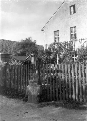 Steinkreuz : Milkel, Steinkreuz mit kurzen Armen und eingeritzter Saufeder vor einem Zaun an der Straßenkreuzung an der Kirche
