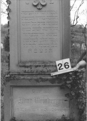 Grabstein 26 (Index: 99805082) Material: Sandstein Zustand: leicht verwittert Maße HxBxT: 207x58x26 cm Beschreibung: Stele auf Doppelsockel mit Rundbogen, Inschriftennische rechteckig mit zwei Kleeblattbögen als Verzierung, Inschriftennische im Sockel querrechteckig Symbolik: Engel im Rundbogen, Blattornamentik am Rundbogen Besonderheit: kulturhistorisch wertvoll / künstl.-formale Gründe: Grabstein 26 (Detailaufnahme)