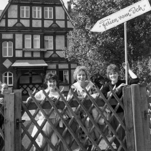 Ferien vom Ich. Drei Feriengäste hinter einem Jägerzaun. Blick zur Ferienunterkunft