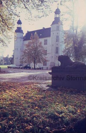 Ahrensburg: Schloss Ahrensburg: Blick auf Nordostfront: davor parkende Pkws und Baum: vorn rechts steinerner Löwe auf Sockel: links Zufahrtsweg: hinten links zwischen Bäumen Turm der Schlosskirche