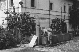 Wohnhaus : Wohnhaus in den Alpen. Eine Frau trinkt aus einem davor liegendem Brunnen.