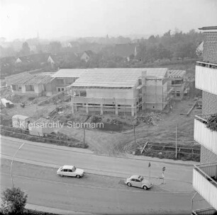 Evangelisches Gemeindehaus: Masurenweg: Rohbau: Blick von einem Hochhaus auf die Baustelle mit Bauwagen, Erdhaufen, Bretterstapel und Baumaterial: vorn zwei Pkw auf Straße: hinten Einfamilienhäuser, Gärten und Bäume