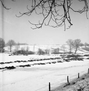 Mühlenbach.: stillgelegter Ausbau: am Ufer Erdhaufen: hinten links Rohre: dahinter Knicklandschaft mit Bäumen und Büschen: vorn rechts Zaun: Winter