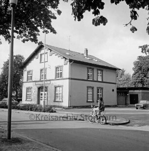 Jugendfreizeitstätte: am Bürgerpark: Einweihung: Außenaufnahme: davor Straße, Verkehrsinsel mit zwei Fahnenmasten, Junge auf Fahrrad, Pkws: rechts Garage: im Vordergrund links Lampenmast: oben überhängende Zweige von Baum