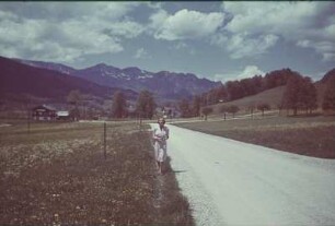 Deutschland. Junge Frau an einer Landstraße vor einer Bergkette (vielleicht in Bayern)