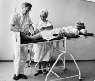 Werbefotografie für einen Prospekt der Fa. Medimex. Eine fahrbare Untersuchungstrage. Ein Arzt untersucht gemeinsam mit einer Schwester eine Patientin