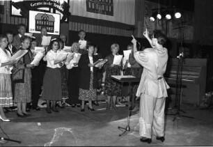 750. Gemeindejubiläum: Abschlussfeier: Sporthalle: Bühne: gemischter Chor beim Singen, mit Dirigentin: Pult, Mikrofone, Klavier, hinten Gemeindeflagge