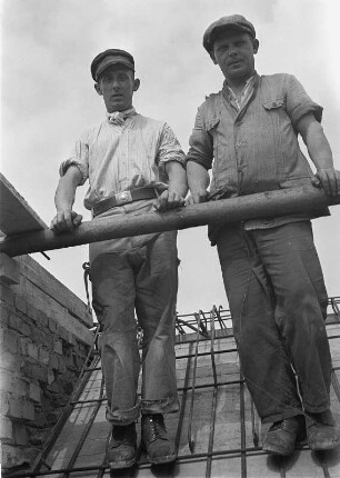 Zwei Bauarbeiter auf einem Dach stehend, an einer Stange angelehnt