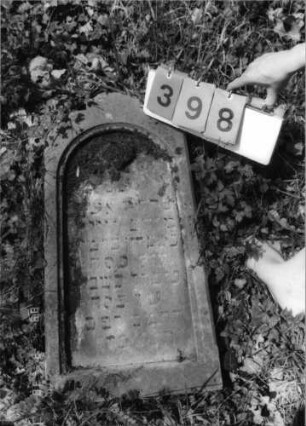Grabstein 398 (Index: 99792066) Material: Sandstein Zustand: verwittert und abgeblättert mit Textverlust, Stele liegend Maße HxBxT: 75x37x15 cm Beschreibung: Stele, Rundbogenabschluß, rechteckig erhöht, Schriftfeld eingetieft Symbolik: Blüte: Grabstein 398