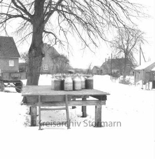 Rethwischdorf: Dorfplatz: links Baum: davor Milchkannenbock: dahinter landwirtschaftlicher Anhänger: im Hintergrund Häuser: Winter