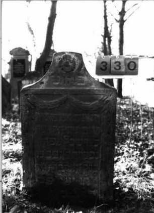 Grabstein 330 (Index: 100115817) Material: Sandstein Zustand: verwittert Maße HxBxT: 95x55x15 cm Beschreibung: Stele mit konkav eingezogenem Segmentbogenabschluß, eingetieftes Schriftfeld Symbolik: das Schriftfeld einrahmende Ranken, Blüte: Grabstein 330