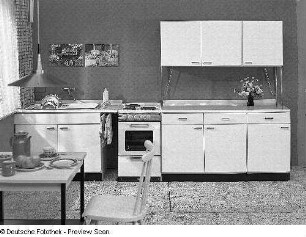 Hängeschrank : Anbauküche (1973). Spüle mit Arbeitsplatte, Elektroherd, Hängeschränken und Schrankteilen der Flachstrecke sowie Küchentisch mit Stuhl
