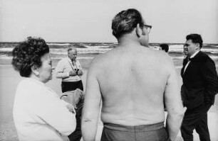 Menschen in unterschiedlichsten Bekleidungsstadien (mit nacktem Oberkörper, mit Anzug und Krawatte) am Strand