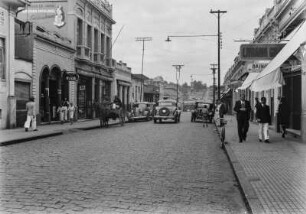 Hauptstraße von Piracicaba : Eine belebte Hauptstraße in Piracicaba. Rechts und links Häusern mit Geschäften, davor Bürgersteige mit Passangten. Auf der Straße Autos und eine Kutsche.