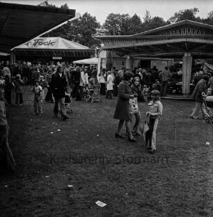 Frühjahrsmarkt: Volksfest: Exer: hinten rechts Karussell, hinten links Jahrmarktsbude: dazwischen Besucher: hinten Mitte Bäume