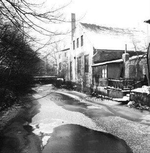 Trave: Stadtarm hinter Hindenburgstraße mit Eisdecke: links Sträucher und Bäume: rechts Gebäude: hinten links Fußgängerbrücke: Winter