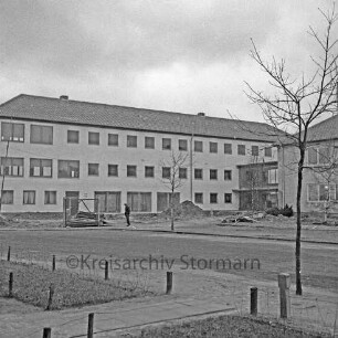 Ahrensburg: Hermann-Löns-Straße: Erweiterungsbau: vor Fertigstellung: rechts Verbindungstrakt, Altbau: auf Vorplatz Baustelle, Erdhaufen, Gerüstteile, Mann: im Vordergrund Straße mit Grünstreifen, Gehweg