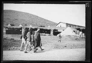 Nador (Marokko): 3 Offiziere [des spanischen 8. Kavallerie-Regiments "Lusitania"] in hellgestreiften Uniformhosen, dunklen Uniformjacken und Tschakos (=militärische Kopfbedeckung in zylindrischer oder konischer Form) bei einem Einheimischen, im Hintergrund ein überdachter Pferdeunterstand, Zelte und ein Gebäude ("Gefangener Mörder auf dem Transport von Nador nach Melilla")