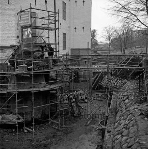 Restaurierung: Sanierung Mauerwerk an Turmfuß: Arbeiter auf Gerüst, Schubkarren: innerer Graben: neue Grabenversteifung mit Holzbohlen am Grabenfuß: Grabenböschung nach Wiederherstellung mit alten Findlingen: im Hintergrund Lübecker Straße, Bauernbrücke, Kastanienallee