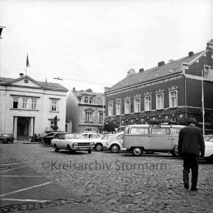 Markt: vorne parkende Pkws, im Hintergrund links das Rathaus, rechts das Stadthaus: Blick von Südosten: