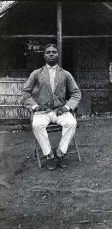 Auf einem Stuhl posierender afrikanischer Mann in Jackett, Hemd und Hose