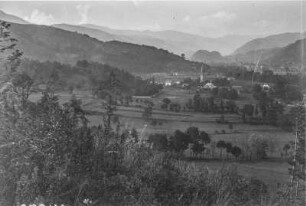 Dorf : Blick auf ein Dorf in einer Hochebene. Im Hintergrund bewaldete Berghänge und eine Kirche, im Vordergrund Felder und Weiden