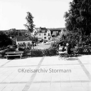 Terrasse in Grünanlage zwischen Marktplatz und Ahrensböker Straße: zwei Sitzbänke, auf einer sitzt Frau, mit Kinderwagen: dahinter Jägerzaun, Rasen, Büsche, Bäume, Fußweg: hinten Häuser an Ahrensböker Straße